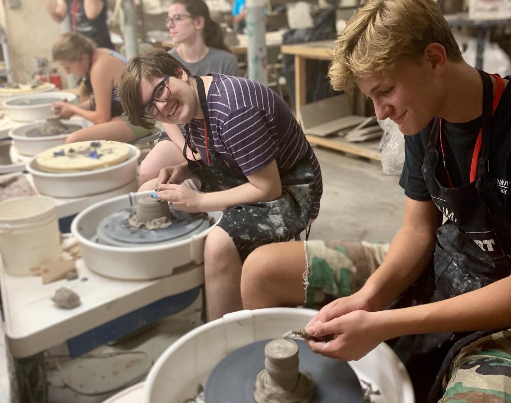 Four teens are sitting at a row of pottery wheels. They all have clay in front of them. One of them is looking and smiling at the camera. 