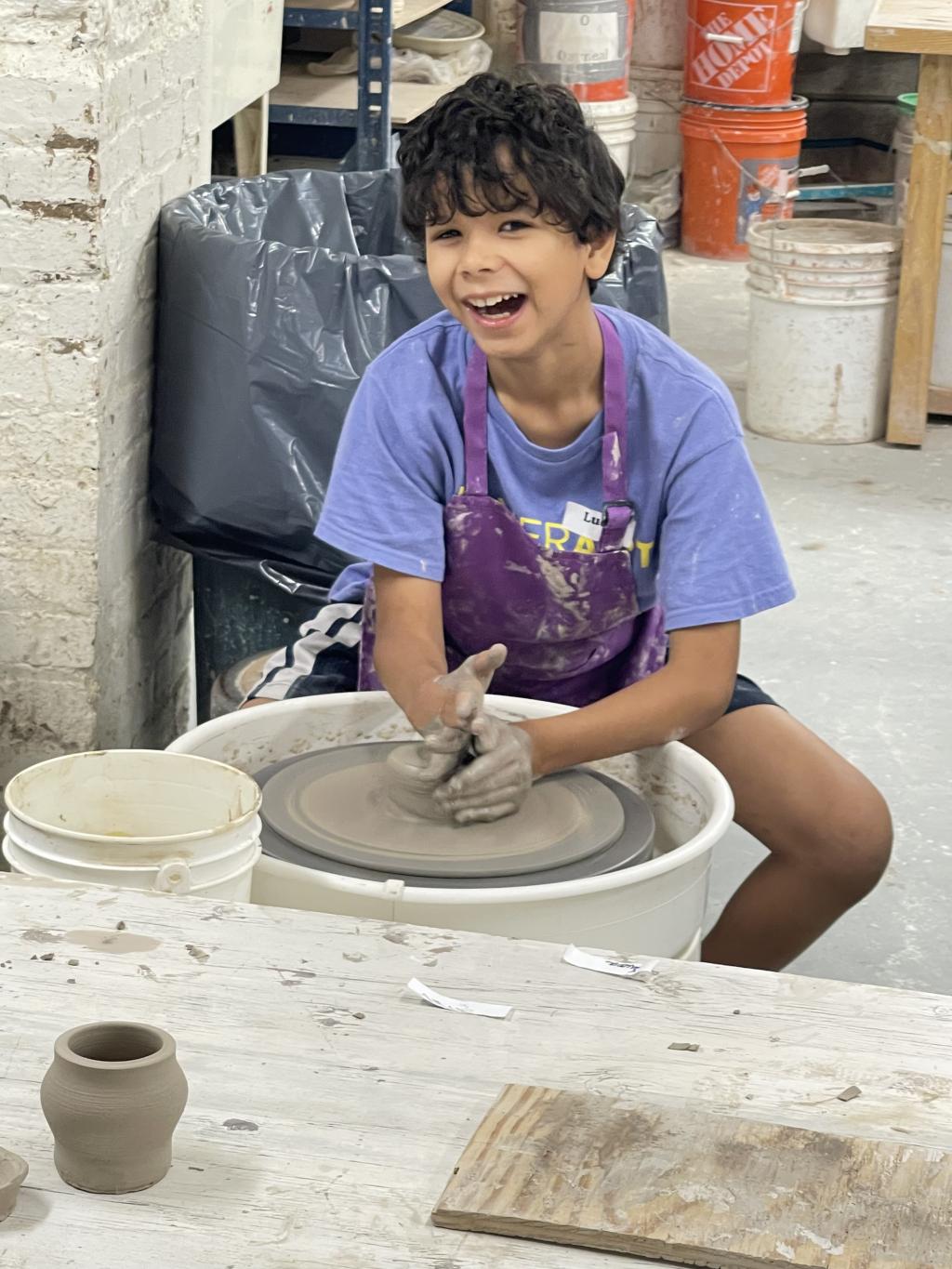 Boy sitting at a potters wheel and smiling wide at the camera
